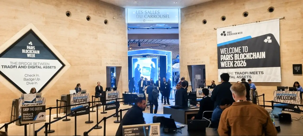 A photograph capturing the main entrance area of the Paris Blockchain Week event at Les Salles du Carrousel in Paris. Numerous attendees are queuing and registering, surrounded by event branding. Signs prominently display the text "PARIS BLOCKCHAIN WEEK, THE BRIDGE BETWEEN TRADFI AND DIGITAL ASSETS"