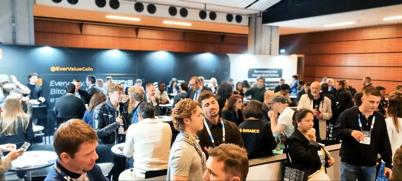 A busy networking lounge at Paris Blockchain Week, filled with attendees talking and connecting. Background displays feature the EverValue project and a Binance logo on a participant's shirt.