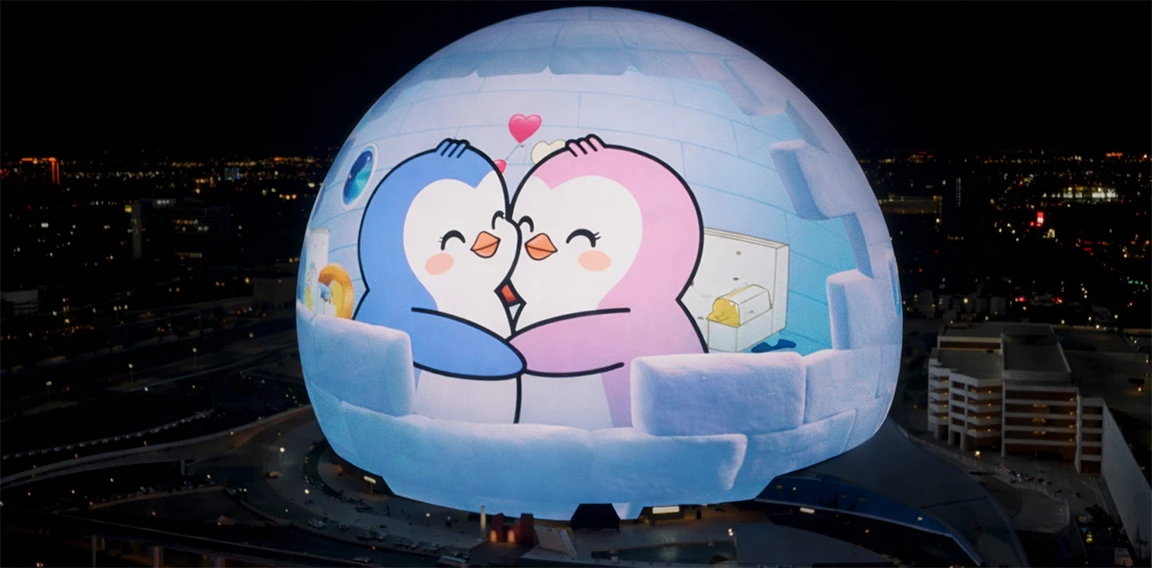 A massive spherical LED dome in a city at night displaying a cartoon of two penguins, one blue and one pink, hugging inside an igloo with small hearts above them.