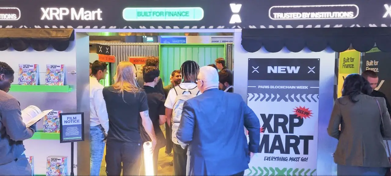 XRP Mart booth at the Paris Blockchain Week event. Numerous attendees are milling around the booth area. Above the stand, a sign reads "XRP Mart" alongside two illuminated signs